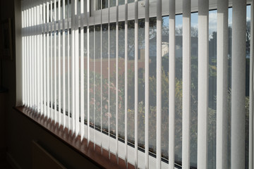 White vertical blinds slats hanging in front of double glazed white frame window. The slats have no cords at the bottom. The focus is shallow.
