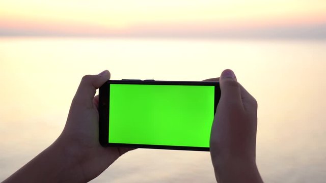 Closeup view of two kid hands holding modern mobile telephone with emty blank green screen. Boy standing isolated at blurry sea water background of golden sunset or sunrise beach. 4k video footage