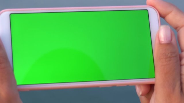 Closeup top view of two female hands holding modern mobile telephone with emty blank green screen. Woman standing at beach isolated at blurry blue sea water background. Real time full hd video footage