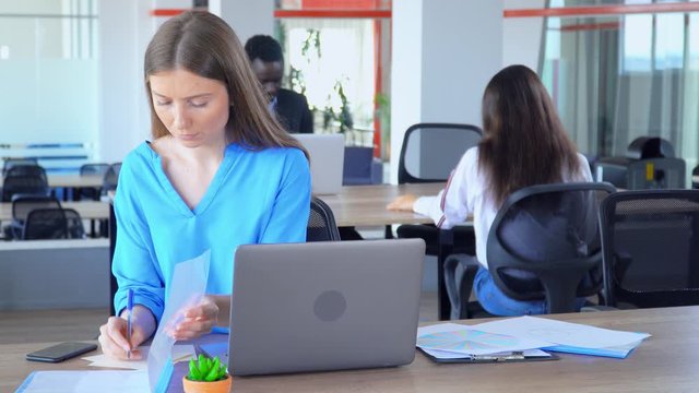 multitasking girl office employee with blond hair at workplace busy young businesswoman in casual blue shirt sitting at desk with chart,tables handwriting notes