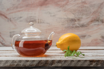 Glass teapot with tea on whitish boards with lemon and a sprig of fresh mint