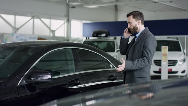 Professional car salesman is telling interested buyer about luxurious car in motor show while man istalkinng with dealer on his cell phone. They are discussing details of the sold car standing at the