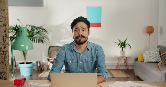 Cute sorrowful Hispanic man feeling sad in front of computer sighs heavily in living room, melancholy bearded adult guy looking to camera dressed in casual blue jeans shirt leaning on hands at table