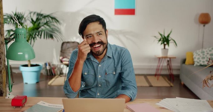 Joyful Hispanic male dreamily looking away from laptop screen touching mustache at home, pleasant brunette young man smiling using gray computer on desk to stay always online in social media network