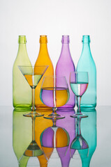 Coloured glass bottles with glasses with water coloured reflections. Reflected on a mirror on a white background