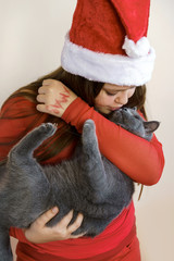 A teenager girl in a red fluffy Santa Claus hat and red sweater holding a fluffy gray British kitten in her arms and smiling. Christmas holiday hugging concept. New year cozy party
