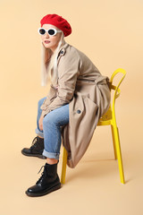 Fashionable mature woman sitting on chair against color background