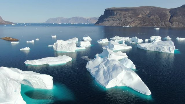 Slow and majestic drone aerial view over white and turquoise Icebergs floating in the calm Baffin Sea, Greenland, with rocky lands sitting in the background of this peaceful arctic landscape