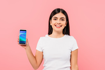  smiling pretty brunette girl holding smartphone with booking app isolated on pink