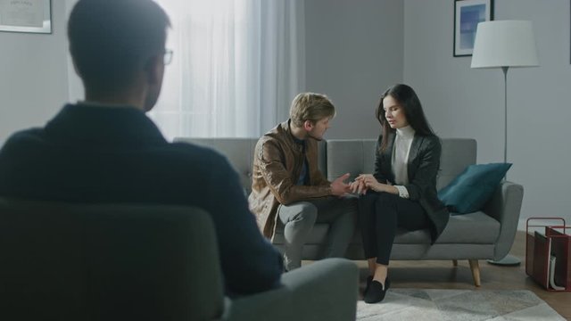 
Young Couple on a Counseling Session with Psychotherapist. Back View of Therapist Taking Notes: Young People Sitting on the Couch, Touching Hands, Discussing Relationship Problems, Finding Solution