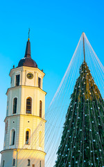 Fototapeta premium Night Cityscape with Christmas Market at Cathedral square in Winter Vilnius, Lithuania. Advent Fair Decoration and Stalls with Crafts Items on the Bazaar. Lithuanian street Xmas and holiday