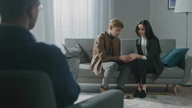 Young Couple on a Counseling Session with Psychotherapist. Back View of Therapist: Tender Partners Sitting on the Couch, Holding Hands, Making up after Quarrel. Husband Kisses Wife&rsquo;s Hand Lovingly