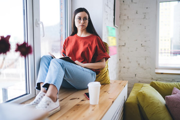Pensive woman writing in notepad sitting on windowsill