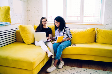 Cheerful multiracial women sitting with laptop on yellow sofa