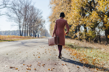 Young redhead lady woman in pink vintage coat and hat with suitcase in retro style walking away along a park road with golden yellow autumnal trees. Outdoor autumn garden relaxation, travel concept