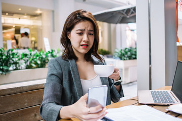 Bewildered young Asian woman with cup using smartphone