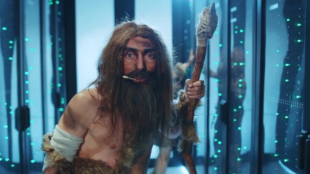Wild caveman of neanderthals tribe chewing animal bone holding a stone-tipped ancient weapon exploring new things in futuristic server data center. Ancient time traveler.
