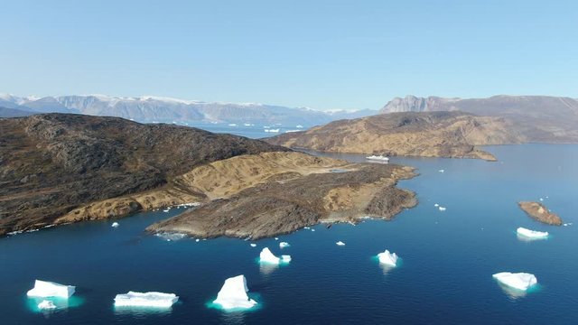 Sliding aerial drone view of Akulleq island, Greenland, with white and turquoise Icebergs floating in the calm Baffin Sea, front facing a rocky, desertic land with several islands