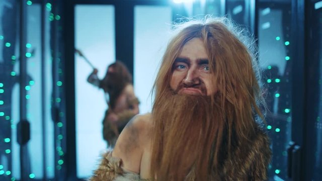 Dangerous caveman leader of hunter-gatherers tribe looking fiercely at camera, threatening with ancient weapon, staying in modern data center. Neanderthals in database.