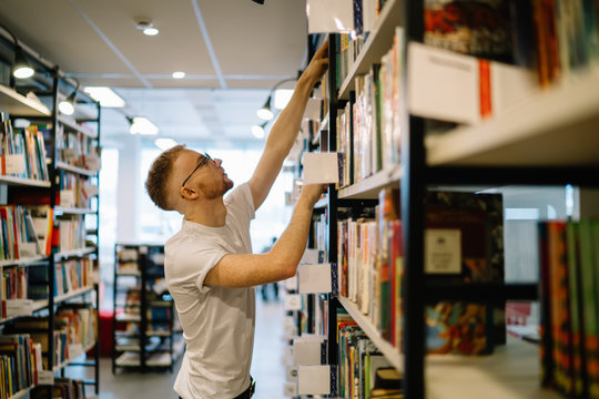 Focused Guy Reaching For Book On Top Shelf