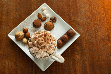 A cup of hot drink with marshmallows, nuts, chocolate on wooden table, blurred background