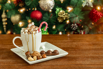 A cup of hot drink with marshmallows, candy stick, nuts, chocolate and caramel on wooden table, Christmas tree background with ornaments and lights, copy space