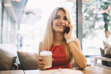 Cheerful female with drink to go speaking on smartphone in cafe