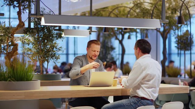 Lockdown of two middle aged businessmen meeting in coffee shop and greeting each other with handshake. Men sitting at counter, looking at laptop screen and discussing business data over coffee