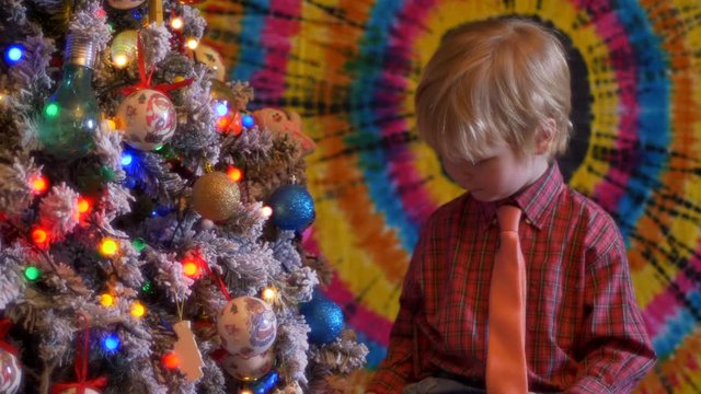 Cute blond boy in plaid shirt and tie in big sunglasses sits next to Christmas tree on colored background. glasses reflect flashing garlands with bright lights. Shallow focus with old movie effect