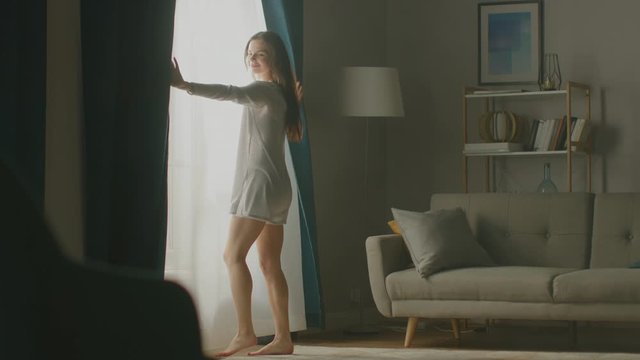 Beautiful Young Brunette Opens Curtains To let Morning Sunlight In, Dances Happily across Cozy Living Room. Side View Of Attractive Girl Rising Early and Greeting New Day. Slow Motion