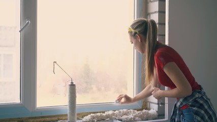 pretty girl with ponytail cleans frame of new window near spray foam insulation pile at construction in empty apartment
