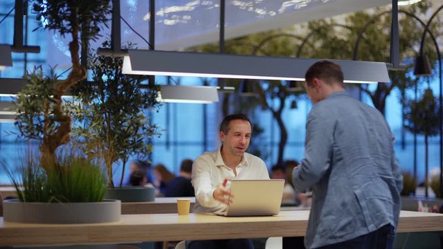 Tracking shot of middle aged man sitting at counter in coffee shop and waiting for business partner. Two businessmen meeting, shaking hands, watching and discussing business presentation on laptop