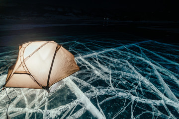 Fire on ice at night. Campground on ice. Tent stands next to bonfire. Lake Baikal. Nearby there is car. Shelter tent and ice are illuminated from the inside. Beautiful bonfire on real cracked ice © ivandanru