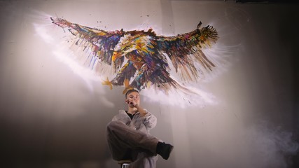 Artist designer draws an eagle on the wall. Craftsman decorator paints a picture with acrylic oil color. Vaper smokes vape. Painter painter dressed in a paint coat. Indoor. Dark magic cinematic look.