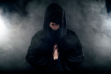 Man dressed in a dark robe looking like a cult leader on a smoky or foggy background.  He looks...
