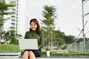 Fototapeta premium Portrait of busy sales woman sitting at business park and using her laptop while writing text on mobile. Businesswoman working online.