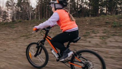 Naklejka premium One caucasian children rides bike road track in dirt park. Girl riding black orange cycle in racetrack. Kid goes do bicycle sports. Biker motion ride with backpack and helmet. Mountain bike hardtail.