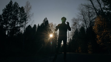 Training an athlete on the roller skaters. Biathlon ride on the roller skis with ski poles, in the helmet. Beautiful sunset silhouette. Autumn workout. Roller sport. Adult man riding on skates.