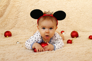 Cute little girl with mouse ears is lying on a beige plaid and playing with red shiny Christmas balls.
