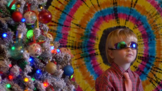 Cute blond boy in plaid shirt and tie in big sunglasses sits next to Christmas tree on colored background. glasses reflect flashing garlands with bright lights. Shallow focus with old movie effect