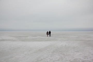 A couple a man and a woman silhouette walk together in the distance on the ice of a frozen snow covered endless winter lake to horizon