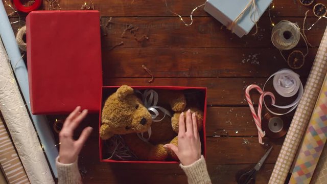Top view of female hands packing plush bear toy into red gift box filled with multicolor shredded tissue paper and decorating it with white ribbon and candy cane while preparing Christmas present