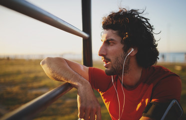 Side view of an exhausted young male athlete listening to music on earphones leaning on bars in gym...