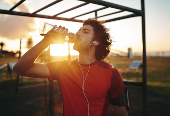 Fitness young athlete man with earphones in his ears drinking water from a reusable bottle after...