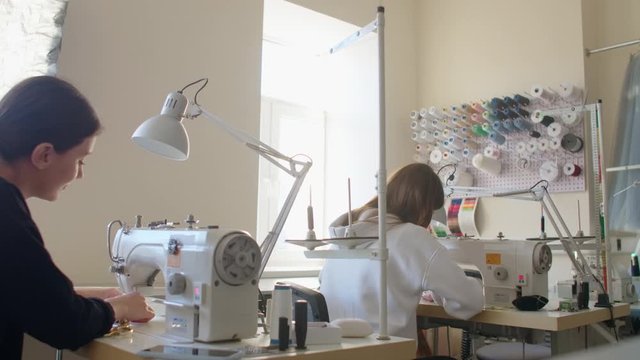 Two women seamstresses working on sewing machines in the tailoring workshop of the garment. The creation of designer dresses. Small business. Two employees work on sewing machines from the back.