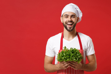 Laughing young bearded male chef cook or baker man in striped apron white t-shirt toque chefs hat isolated on red background. Cooking food concept. Mock up copy space. Hold green fresh salad leaves.