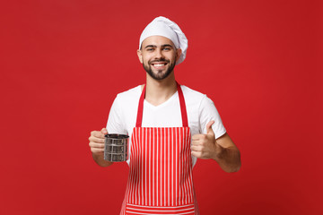 Smiling male chef cook or baker man in striped apron toque chefs hat posing isolated on red background. Cooking food concept. Mock up copy space. Hold metal iron sieve sifting flour, showing thumb up.