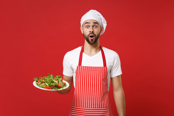 Shocked young bearded male chef cook or baker man in striped apron toque chefs hat posing isolated on red wall background. Cooking food concept. Mock up copy space. Hold plate with vegetable salad.