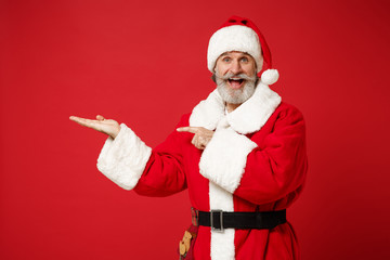 Fototapeta na wymiar Excited elderly gray-haired mustache bearded Santa man in Christmas hat posing isolated on red background. Happy New Year 2020 celebration holiday concept. Mock up copy space. Point finger hand aside.