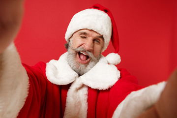 Close up of elderly gray-haired mustache bearded Santa man in Christmas hat posing isolated on red background. New Year 2020 celebration concept. Mock up copy space. Doing selfie shot on mobile phone.